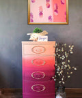 Dixie Belle Paint Plum Crazy Chalk Mineral Paint is the perfect paint for any DIY project! End table chalk painted in maroon, coral and apricot blended together with gold on the trims in the front. Staged with a conch shell with a plant, a pineapple picture and glass vase with a plant inside.
