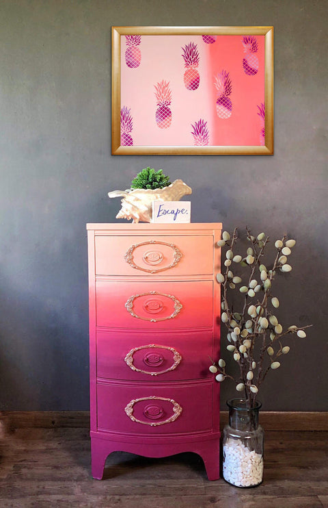 Dixie Belle Paint Plum Crazy Chalk Mineral Paint is the perfect paint for any DIY project! End table chalk painted in maroon, coral and apricot blended together with gold on the trims in the front. Staged with a conch shell with a plant, a pineapple picture and glass vase with a plant inside.