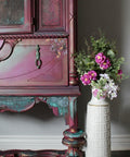 Dixie Belle Paint Plum Crazy Chalk Mineral Paint is the perfect paint for any DIY project! Up close of a display cabinet chalk painted in a maroon color with patina to give it a rusty look and a floral rub on transfer on it. Staged next to a white vase with pink flowers.