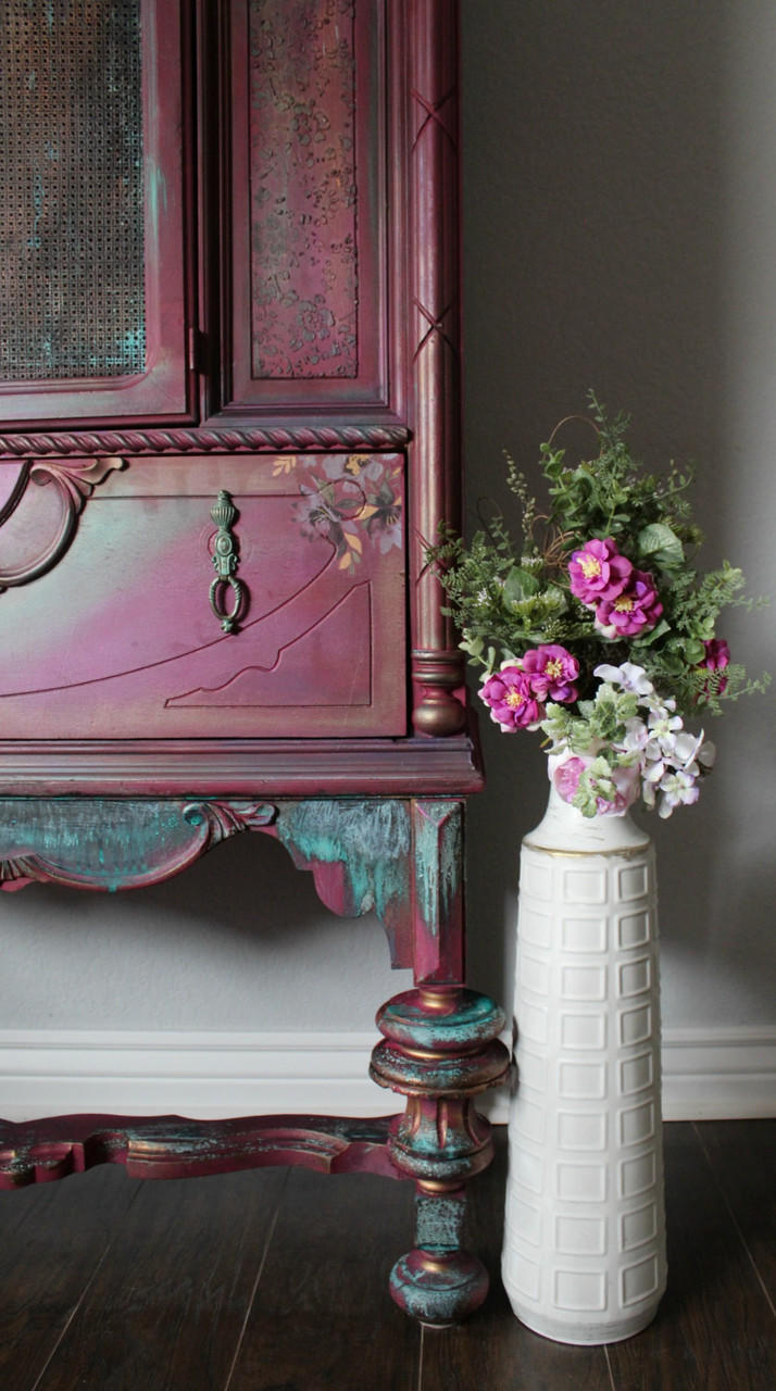 Dixie Belle Paint Plum Crazy Chalk Mineral Paint is the perfect paint for any DIY project! Up close of a display cabinet chalk painted in a maroon color with patina to give it a rusty look and a floral rub on transfer on it. Staged next to a white vase with pink flowers.