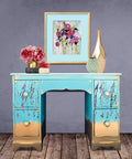 Dixie Belle Paint Pure Ocean Chalk Mineral Paint is the perfect paint for any DIY project! Blue chalk painted desk with gold ends and hardware and floral rub on transfers. Staged with pink plants and gold vases.