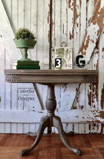 Dixie Belle Paint Putty Chalk Mineral Paint is the perfect paint for any DIY project! A muted taupe colored table staged in front of a rustic door with various green props on top such as books, plant with a plant stand and the letter G.