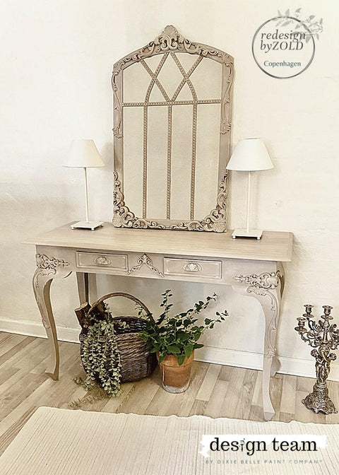 Dixie Belle Paint Putty Chalk Mineral Paint is the perfect paint for any DIY project! A muted taupe vanity with a decorative mirror on the top surrounded by various staging props such as 2 white lamps staged on both sides of vanity, a basket with greenery underneath the vanity, a brown plant pot with greenery underneath the vanity also, and a metal candelabra on the right of the vanity.