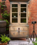 Dixie Belle Paint Putty Chalk Mineral Paint is the perfect paint for any DIY project! An armoire cabinet painted in a muted taupe with various brown tones and a steampunk transfer on the front staged in front of a brick building with greenery surrounding.