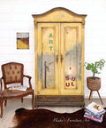 Dixie Belle Paint Rebel Yell-ow Chalk Mineral Paint is the perfect paint for any DIY project! An armoire painted in this vintage yellow all over and accented on the top and bottom trim with brown. The front shows gray in the middle with the words "art feed your soul" in an artistic manner. This armoire is staged with brown furniture, a plant stand on the right, a wooden brown table in front and a brown antique chair to the left.