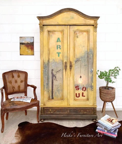 Dixie Belle Paint Rebel Yell-ow Chalk Mineral Paint is the perfect paint for any DIY project! An armoire painted in this vintage yellow all over and accented on the top and bottom trim with brown. The front shows gray in the middle with the words "art feed your soul" in an artistic manner. This armoire is staged with brown furniture, a plant stand on the right, a wooden brown table in front and a brown antique chair to the left.