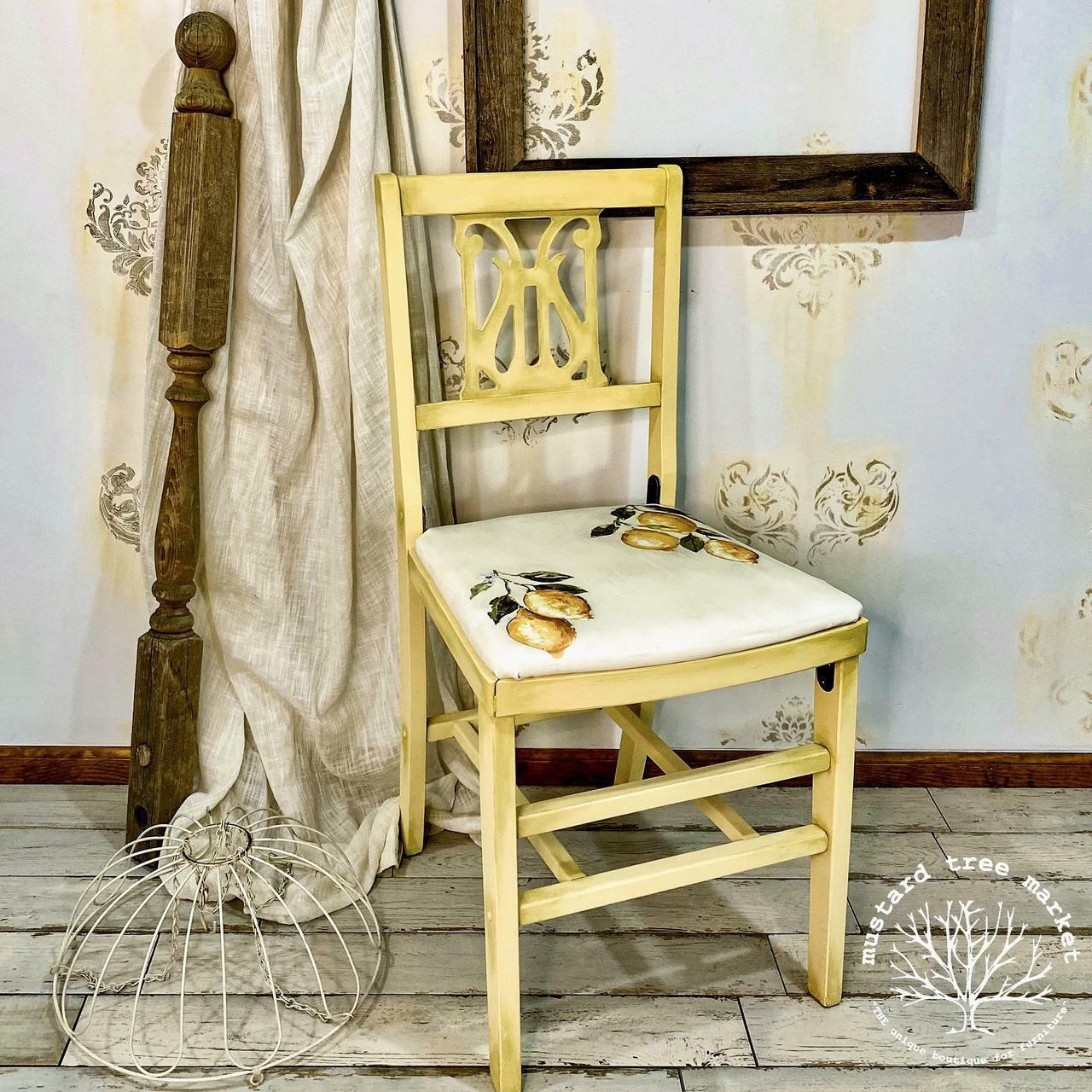 Dixie Belle Paint Rebel Yell-ow Chalk Mineral Paint is the perfect paint for any DIY project! A kitchen chair with this vintage yellow color all over the frame with a white seat. The white seat has a lemon transfer on top and it is staged with various household items.
