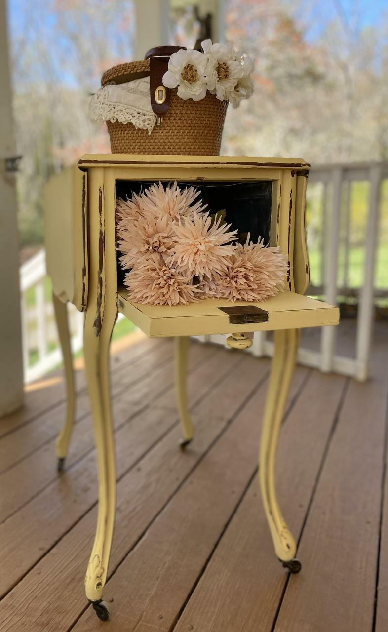 Dixie Belle Paint Rebel Yell-ow Chalk Mineral Paint is the perfect paint for any DIY project! This folding end table on wheels is sporting this vintage yellow throughout with a bit of distressing along the trim. This side table is staged on a porch and has the back open up into one opening with flowers and a basket on top of the end table.