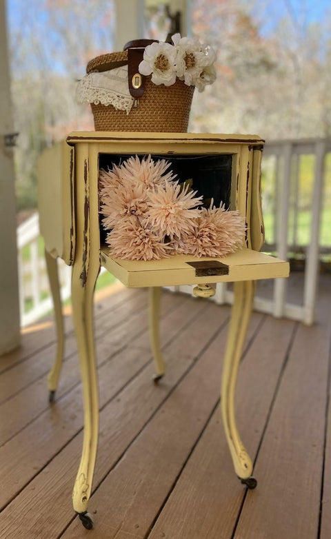Dixie Belle Paint Rebel Yell-ow Chalk Mineral Paint is the perfect paint for any DIY project! This folding end table on wheels is sporting this vintage yellow throughout with a bit of distressing along the trim. This side table is staged on a porch and has the back open up into one opening with flowers and a basket on top of the end table.
