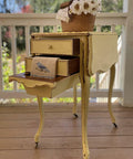 Dixie Belle Paint Rebel Yell-ow Chalk Mineral Paint is the perfect paint for any DIY project! This folding end table on wheels is sporting this vintage yellow throughout with a bit of distressing along the trim. This side table is staged on a porch and has a hand towel with a bird draped over the second drawer and a basket with faux flowers on top of the end table.