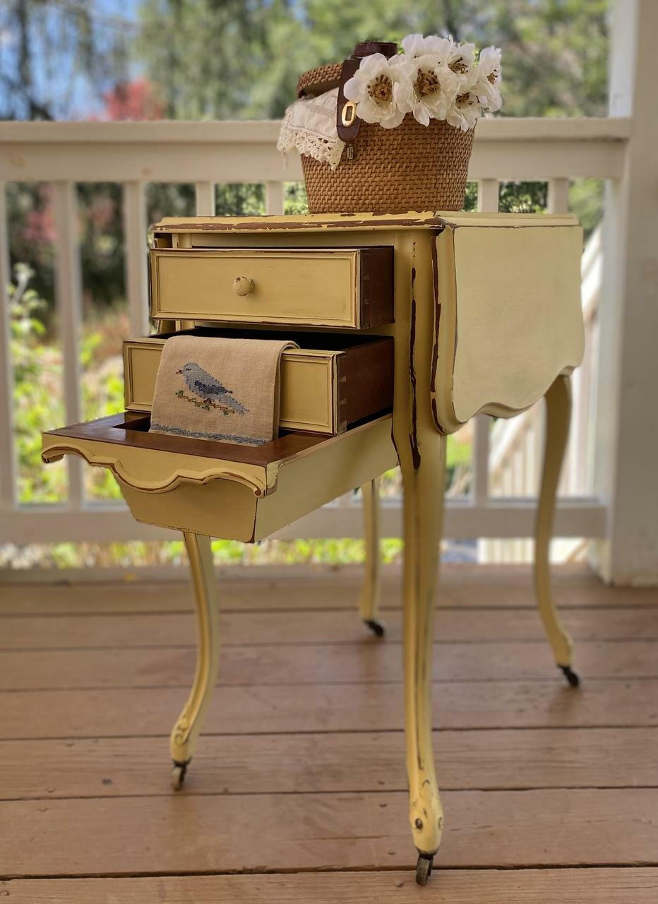 Dixie Belle Paint Rebel Yell-ow Chalk Mineral Paint is the perfect paint for any DIY project! This folding end table on wheels is sporting this vintage yellow throughout with a bit of distressing along the trim. This side table is staged on a porch and has a hand towel with a bird draped over the second drawer and a basket with faux flowers on top of the end table.