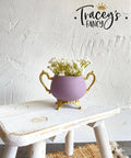 Purple chalk painted pitcher with gold legs and handles. Staged with white flowers inside, on a white painted distressed stool.