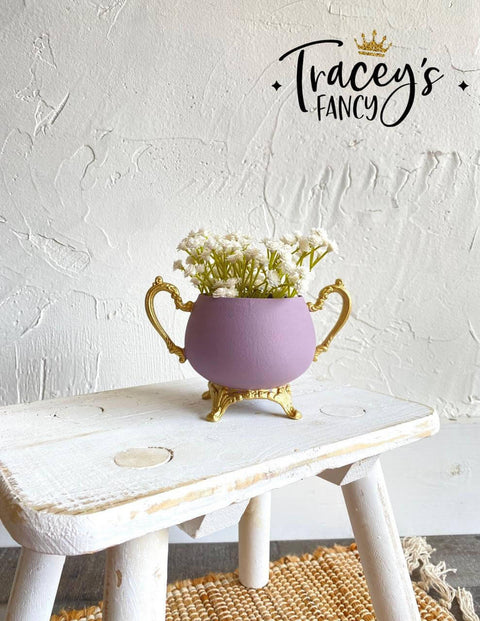 Purple chalk painted pitcher with gold legs and handles. Staged with white flowers inside, on a white painted distressed stool.