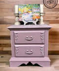 Purple chalk painted nightstand with scrolls on the sides of the drawers. Staged with two large candlesticks and a colorful painting on top.