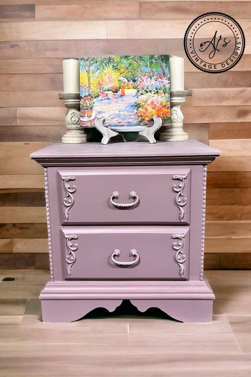 Purple chalk painted nightstand with scrolls on the sides of the drawers. Staged with two large candlesticks and a colorful painting on top.