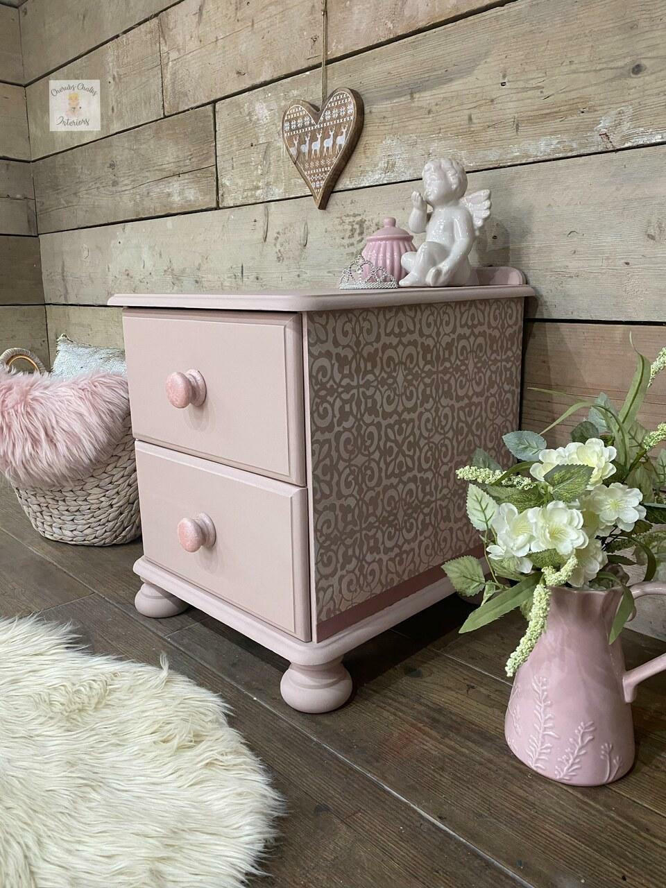 Dixie Belle Paint Soft Pink Chalk Mineral Paint is the perfect paint for any DIY project! A small side table with 2 drawers painted in the soft pink chalk mineral paint and is accompanied  by a stencil on the sides and is staged with plants on the right and other household decor items.