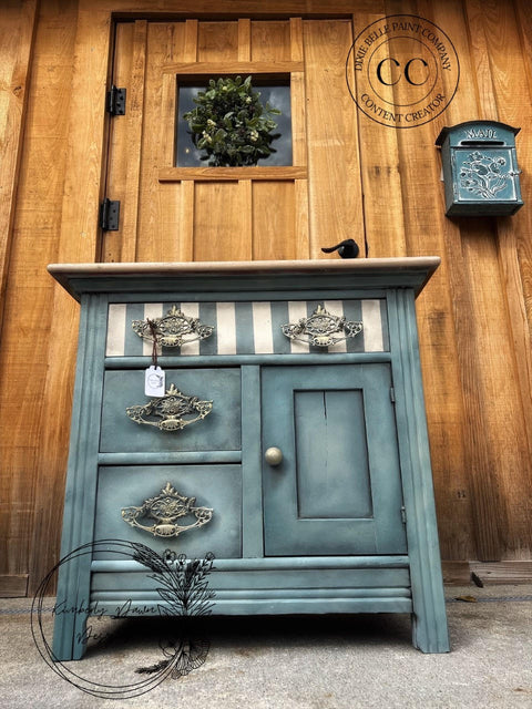 Dixie Belle Paint Stormy Seas Chalk Mineral Paint is the perfect paint for any DIY project!
Vanity cabinet painted in Stormy Seas Chalk Mineral Paint with a striped top drawer and silver hardware. Staged Infront of rustic wood door and wall with a wall mounted mailbox.