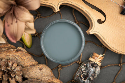 Dixie Belle Paint Stormy Seas Chalk Mineral Paint is the perfect paint for any DIY project!
Top view of open container of Stormy Seas Chalk Mineral Paint on top of a black cover and a rope netting , staged with a violin, flowers and pinecones.