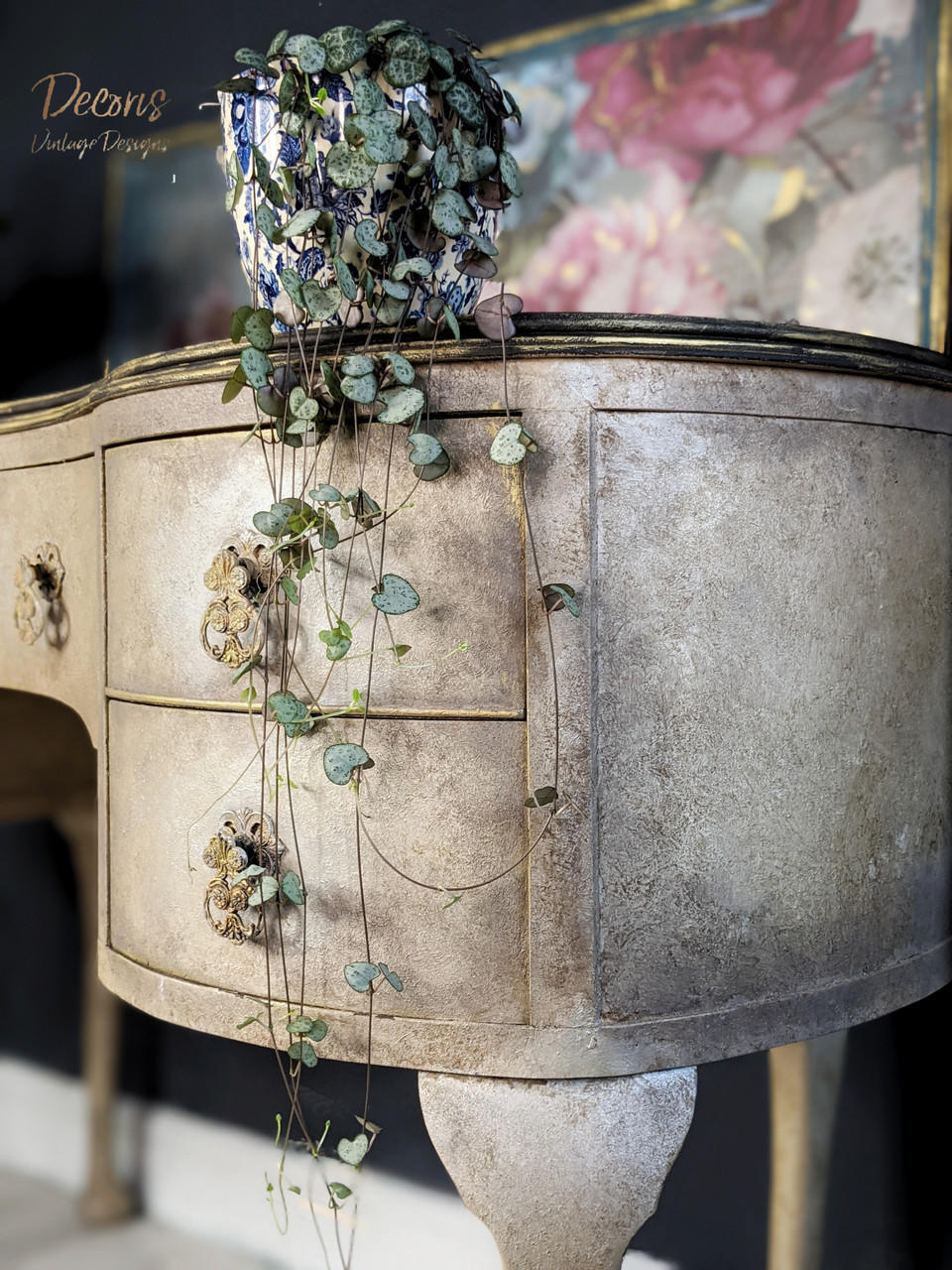Dixie Belle Paint Van Dyke Brown Glaze. The right side of a light brown chalk painted des with darker brown edges and gold handles.  A green plant in a pot sits on top of the desk and a floral picture is in the background.