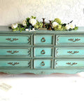 Dixie Belle Paint Vintage Duck Egg Chalk Mineral Paint is the perfect paint for any DIY project!  A greenish blue chalk painted dresser with gold handles sits on a white floor in front of a white wall.  White flowers with greenery and gold candle sticks sit on top of the dresser.  An open book with loose pages sits on the floor to the left of the dresser.