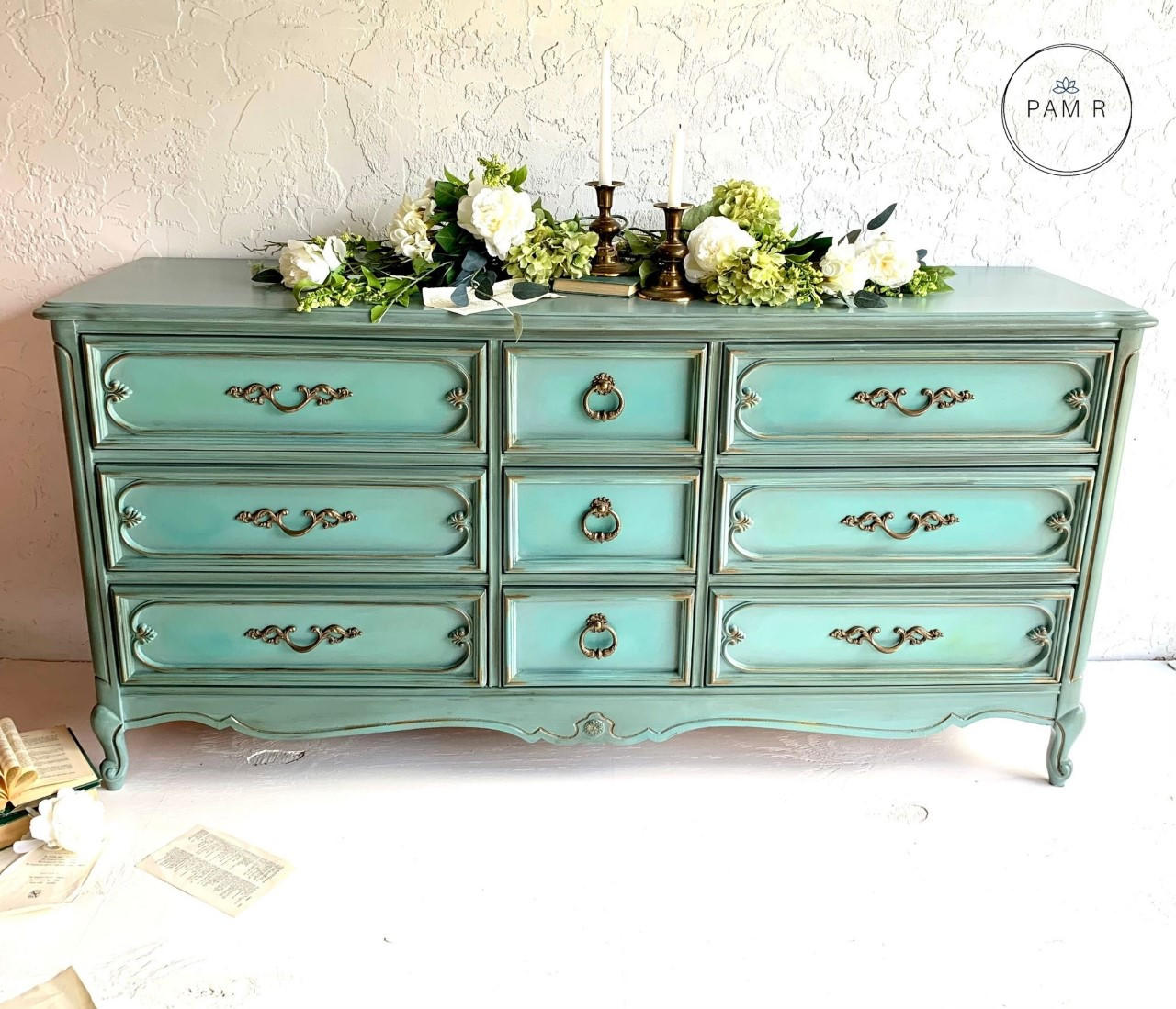 Dixie Belle Paint Vintage Duck Egg Chalk Mineral Paint is the perfect paint for any DIY project!  A greenish blue chalk painted dresser with gold handles sits on a white floor in front of a white wall.  White flowers with greenery and gold candle sticks sit on top of the dresser.  An open book with loose pages sits on the floor to the left of the dresser.