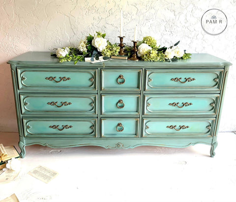 Dixie Belle Paint Vintage Duck Egg Chalk Mineral Paint is the perfect paint for any DIY project!  A greenish blue chalk painted dresser with gold handles sits on a white floor in front of a white wall.  White flowers with greenery and gold candle sticks sit on top of the dresser.  An open book with loose pages sits on the floor to the left of the dresser.