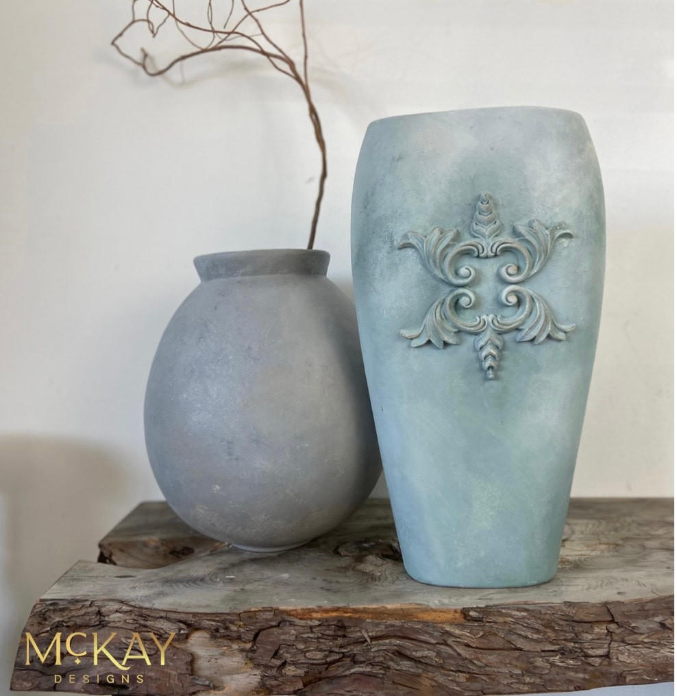 Dixie Belle Paint Vintage Duck Egg Chalk Mineral Paint is the perfect paint for any DIY project!  A small rounded gray chalk painted clay pot and a grayish-blue blended chalk painted clay pot with ornamental decorations sit on a piece of wood, in front of a white wall.  A stick is coming out of the small gray pot.