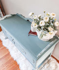 Dixie Belle Paint Vintage Duck Egg Chalk Mineral Paint is the perfect paint for any DIY project!  Top view of a blue chalk painted cabinet sitting on a white furry rug, on top of a wooden floor.  A basket of white flowers and greenery sit on top of the cabinet, next to a white cup and a brown book.