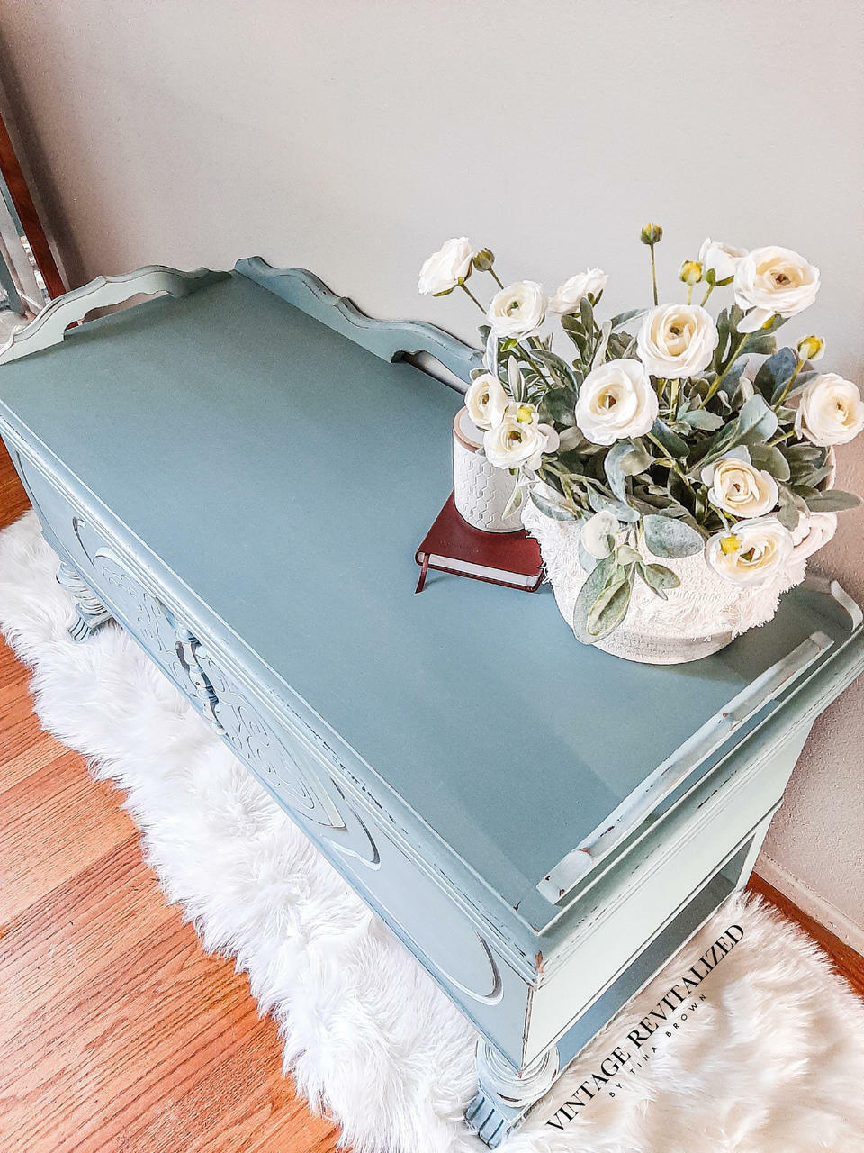 Dixie Belle Paint Vintage Duck Egg Chalk Mineral Paint is the perfect paint for any DIY project!  Top view of a blue chalk painted cabinet sitting on a white furry rug, on top of a wooden floor.  A basket of white flowers and greenery sit on top of the cabinet, next to a white cup and a brown book.