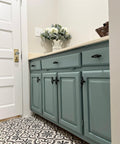 Dixie Belle Paint Vintage Duck Egg Chalk Mineral Paint.  Grayish-blue chalk painted bathroom cabinets with black knobs and a white counter top.  A white chalk painted canister with gray letters on it sits on top of the counter with white flowers and greenery inside, as well as a dark brown woven basket.  The floor of the bathroom has black, gray and white tiles on it and the door is painted white with a gold door knob.