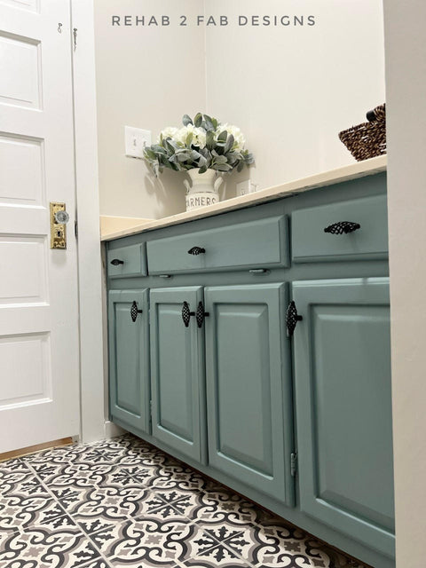 Dixie Belle Paint Vintage Duck Egg Chalk Mineral Paint.  Grayish-blue chalk painted bathroom cabinets with black knobs and a white counter top.  A white chalk painted canister with gray letters on it sits on top of the counter with white flowers and greenery inside, as well as a dark brown woven basket.  The floor of the bathroom has black, gray and white tiles on it and the door is painted white with a gold door knob.