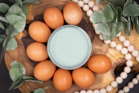 Dixie Belle Paint Vintage Duck Egg Chalk Mineral Paint is the perfect paint for any DIY project!  Top view of an open jar of Vintage Duck Egg Chalk Mineral Paint sitting on a circular piece of wood with brown eggs surrounding it and white beads and green foliage around the eggs.