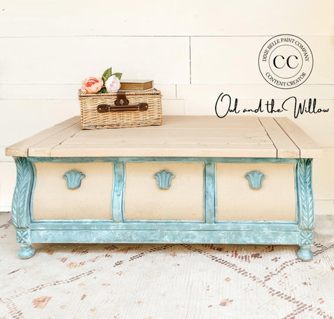 Dixie Belle Paint Vintage Duck Egg Chalk Mineral Paint is the perfect paint for any DIY project!  A square coffee table chalk painted off-white and bluish green sits on a white & brown carpet in front of a white wall.  A tan basket with a dark brown handle sits on top of the table with two light colored flowers and a light colored book with gold pages on top of the basket.