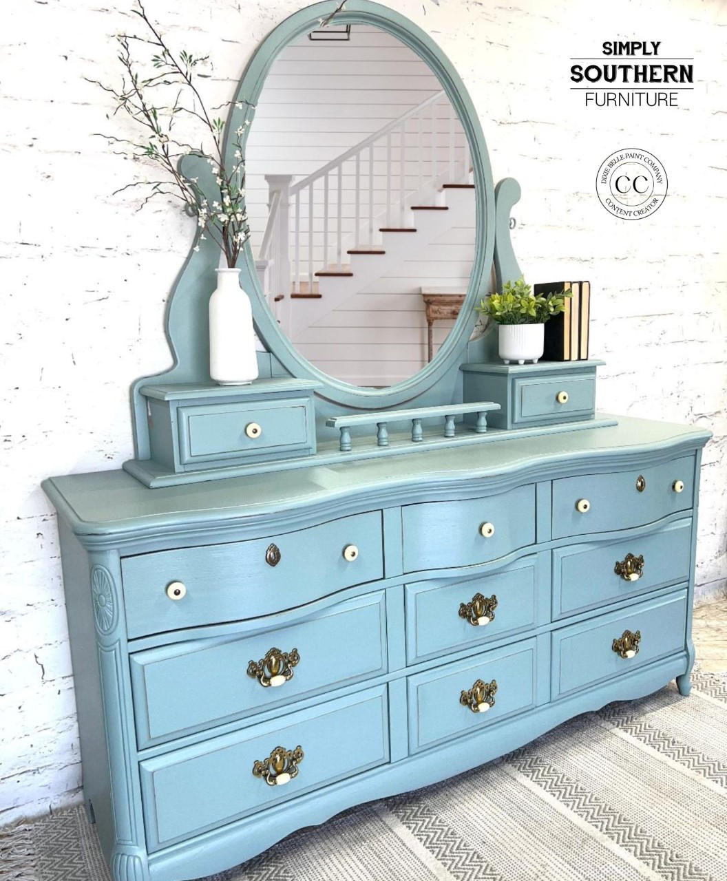 Dixie Belle Paint Vintage Duck Egg Chalk Mineral Paint.  A blue chalk painted dresser with a large oval mirror on top of it and bronze & white handles.  A white vase holding brown sticks with white flowers and greenery sits to the left of the mirror on the dresser.  A white pot holding a green plant and 3 black books sit to the right of the mirror.  The dresser sits on a gray and white striped rug, in front of a white brick wall.