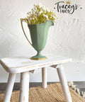 Green chalk painted pitcher with a gold base and handle. Staged with white flowers inside, on a white painted distressed stool.
