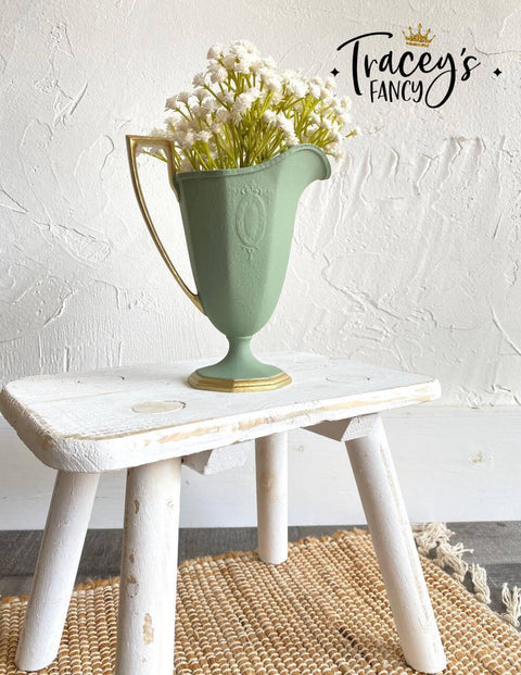 Green chalk painted pitcher with a gold base and handle. Staged with white flowers inside, on a white painted distressed stool.
