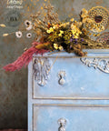 Dixie Belle Paint Whitewash Glaze.  The upper left corner of a blue chalk painted dresser with whitewash applied to the front of it and wooden embellishments.  The dresser is in front of a gray wall with the Victorian Damask stencil chalk painted in gold.  A bunch of flowers in brown, pink, yellow, white and purple sit on top of the dresser.