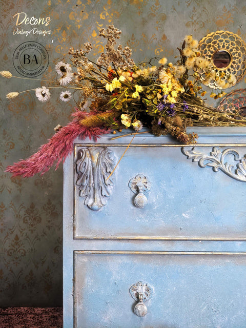 Dixie Belle Paint Whitewash Glaze.  The upper left corner of a blue chalk painted dresser with whitewash applied to the front of it and wooden embellishments.  The dresser is in front of a gray wall with the Victorian Damask stencil chalk painted in gold.  A bunch of flowers in brown, pink, yellow, white and purple sit on top of the dresser.
