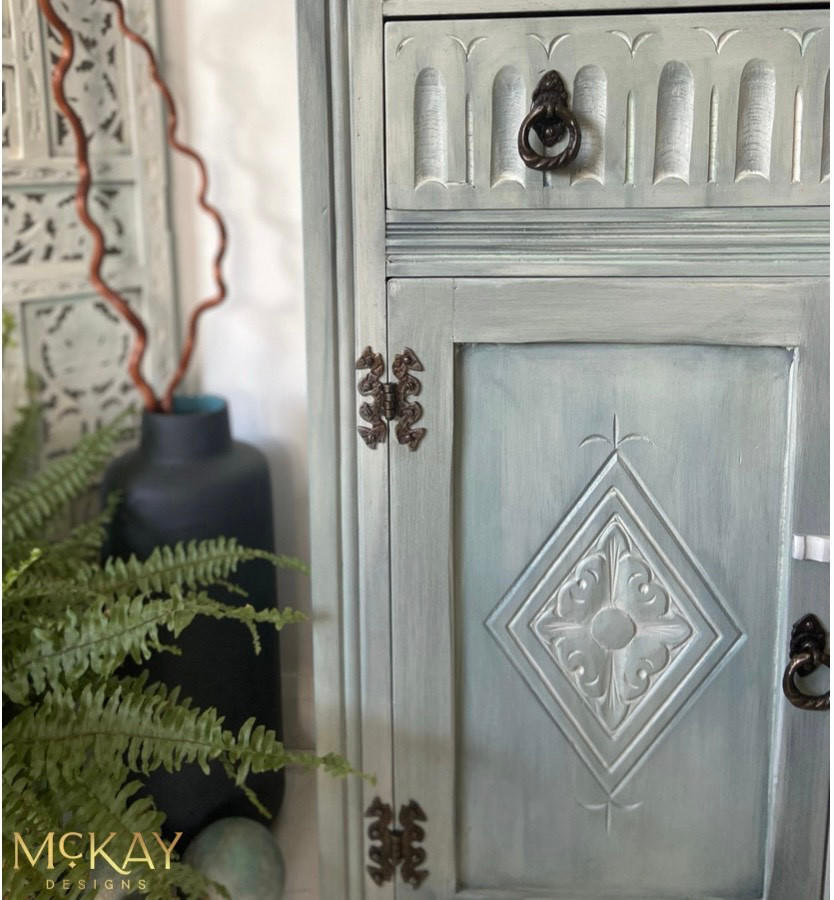 Dixie Belle Paint Whitewash Glaze. A gray chalk painted cabinet with bronze hardware has Whitewash Glaze applied to the front of it.  To the left of the cabinet is a black chalk painted vase holding two brown sticks and a green plant.