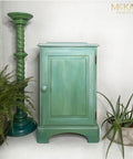 Dixie Belle Paint Whitewash Glaze.  A green chalk painted cabinet that has Whitewash Glaze on the top of the cabinet and coming down the front of the cabinet sitting on a white floor, in front of a white wall.  A green aloe plant is in a gray and white chalk painted pot on the floor to the right of the cabinet.  A two-tone green chalk painted column sits to the right of the cabinet with a green fern sitting inside a brown woven basket next to it.