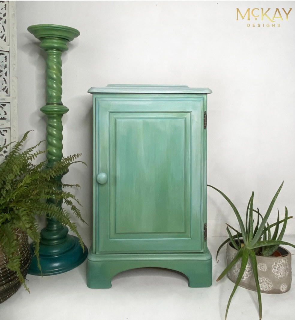 Dixie Belle Paint Whitewash Glaze.  A green chalk painted cabinet that has Whitewash Glaze on the top of the cabinet and coming down the front of the cabinet sitting on a white floor, in front of a white wall.  A green aloe plant is in a gray and white chalk painted pot on the floor to the right of the cabinet.  A two-tone green chalk painted column sits to the right of the cabinet with a green fern sitting inside a brown woven basket next to it.