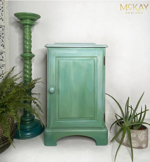 Dixie Belle Paint Whitewash Glaze.  A green chalk painted cabinet that has Whitewash Glaze on the top of the cabinet and coming down the front of the cabinet sitting on a white floor, in front of a white wall.  A green aloe plant is in a gray and white chalk painted pot on the floor to the right of the cabinet.  A two-tone green chalk painted column sits to the right of the cabinet with a green fern sitting inside a brown woven basket next to it.
