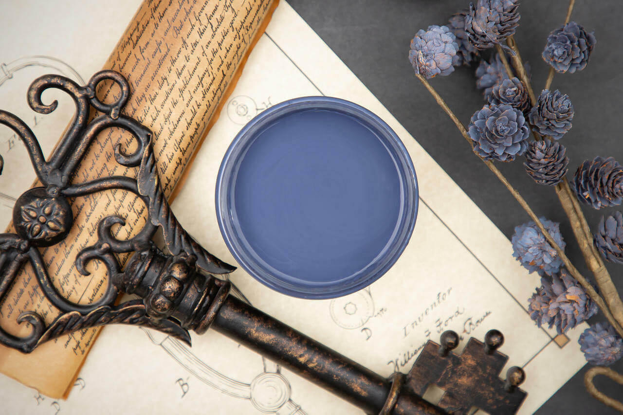 Dixie Belle Paint Yankee Blue Chalk Mineral Paint is the perfect paint for any DIY project!  Top view of an open can of Yankee Blue chalk Mineral Paint on a white sheet of paper with pencil drawings sitting on a black table.  Blue chalk painted mini pinecones sit to the right of the jar on brown sticks.  A bronze antique ke sits on a brown rolled up scroll of paper with handwritten words.
