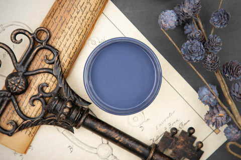 Dixie Belle Paint Yankee Blue Chalk Mineral Paint is the perfect paint for any DIY project!  Top view of an open can of Yankee Blue chalk Mineral Paint on a white sheet of paper with pencil drawings sitting on a black table.  Blue chalk painted mini pinecones sit to the right of the jar on brown sticks.  A bronze antique ke sits on a brown rolled up scroll of paper with handwritten words.
