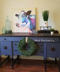 Dixie Belle Paint Yankee Blue Chalk Mineral Paint is the perfect paint for any DIY project!  A Yankee Blue chalk painted desk with black legs and desktop.  A green wreath hangs in front of the desk.  Two clear glass bottles, a multi-colored painting of a cow and green plants in white pots sit on top of the desk.  The desk sits on a brown wooden floor in front of a white wall.