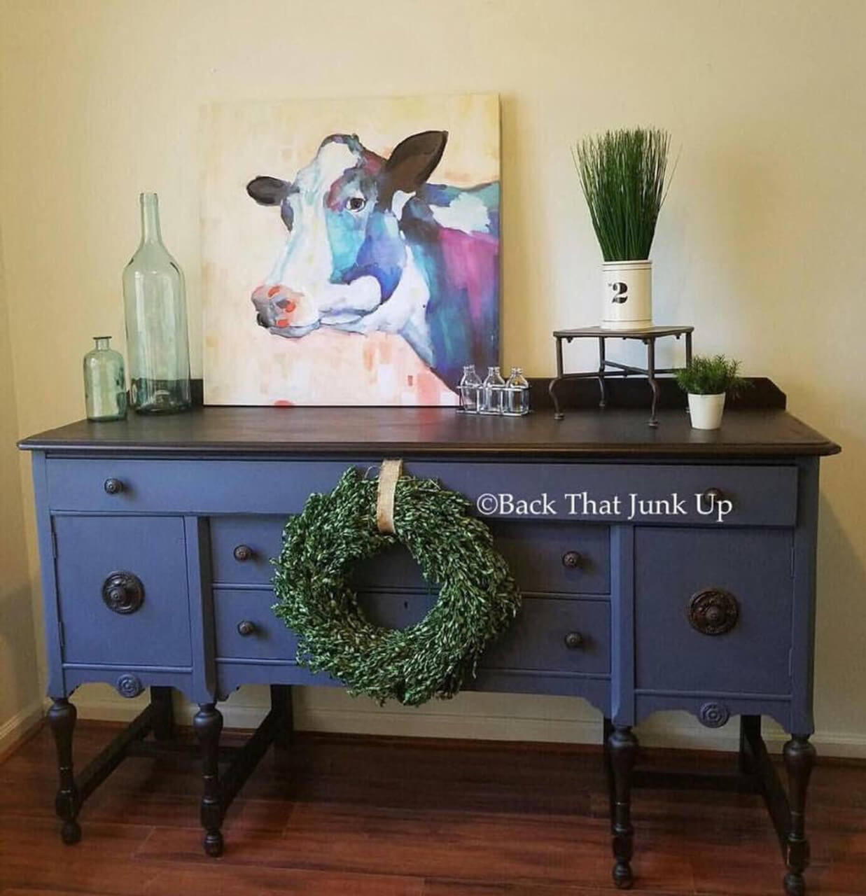 Dixie Belle Paint Yankee Blue Chalk Mineral Paint is the perfect paint for any DIY project!  A Yankee Blue chalk painted desk with black legs and desktop.  A green wreath hangs in front of the desk.  Two clear glass bottles, a multi-colored painting of a cow and green plants in white pots sit on top of the desk.  The desk sits on a brown wooden floor in front of a white wall.