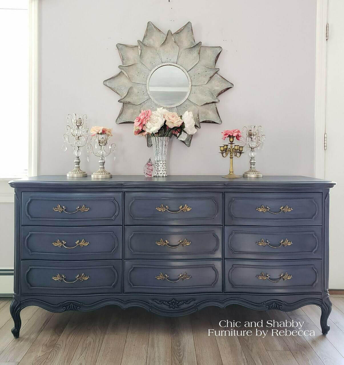 Dixie Belle Paint Yankee Blue Chalk Mineral Paint is the perfect paint for any DIY project!  A Yankee Blue chalk painted dresser with gold hardware sits on a light brown wooden floor, in front of a gray wall with white trim.  A round mirror with silver flower petals hangs on the wall above the dresser.  Several silver candlesticks with clear hanging ornaments sit on top of the dresser, along with pink and white roses inside of a clear glass vase.