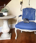 Dixie Belle Paint Yankee Blue Chalk Mineral Paint is the perfect paint for any DIY project!  A white and Yankee Blue chalk painted chair sits on a brown wooden floor in front of a tan wall with white trim.  To the left of the chair is a round chalk painted table with a wire bird cage holding a green plant in a white pot.  A black ornamental piece of decor also sits on the table.