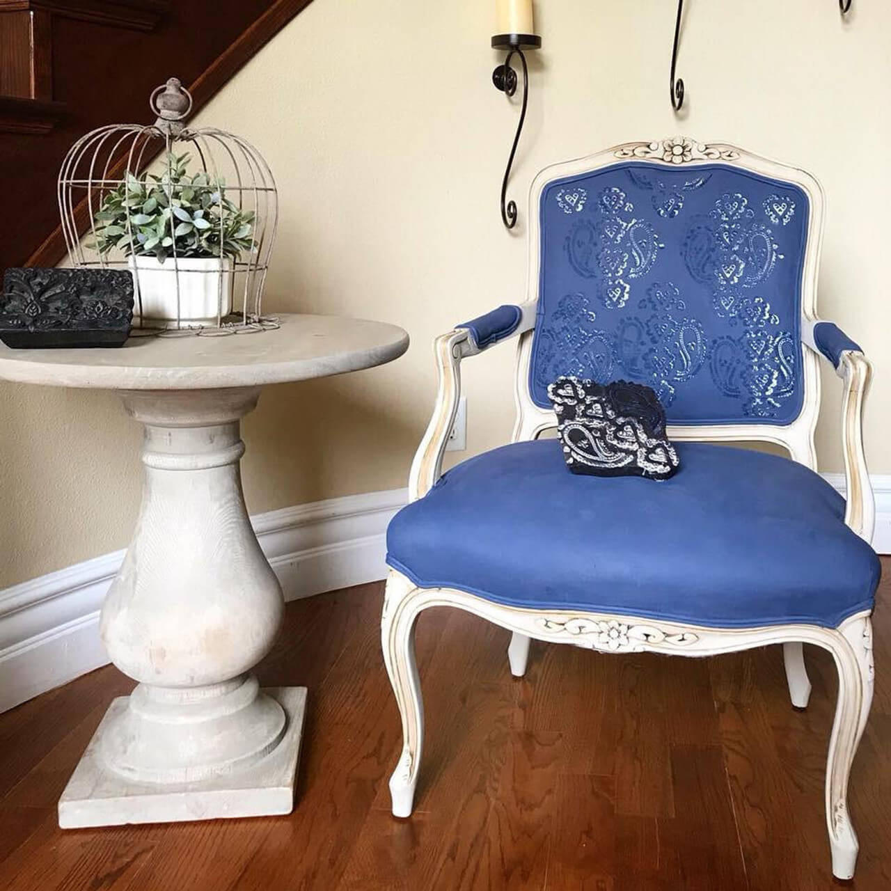 Dixie Belle Paint Yankee Blue Chalk Mineral Paint is the perfect paint for any DIY project!  A white and Yankee Blue chalk painted chair sits on a brown wooden floor in front of a tan wall with white trim.  To the left of the chair is a round chalk painted table with a wire bird cage holding a green plant in a white pot.  A black ornamental piece of decor also sits on the table.