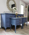 Dixie Belle Paint Yankee Blue Chalk Mineral Paint is the perfect paint for any DIY project!  A Yankee Blue chalk painted dresser and two matching blue chalk painted night stands sit on a light brown floor, in front of a gray wall with white trim.  A round mirror in a silver frame hang on the wall above the blue dresser.  A white and pink flowers, a clear vase, a bronze candlestick and other decorations sit on top of the chalk painted pieces.